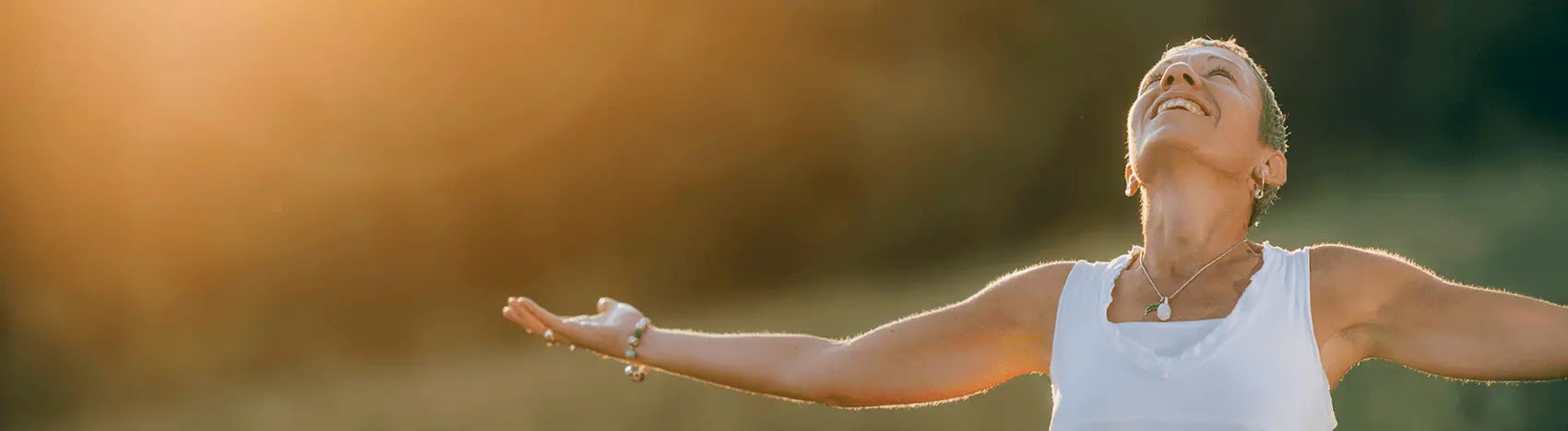 A mature woman stands in a sunlit field with her arms outstretched and a serene smile, embracing the warm golden light. This image represents the joy and freedom of living without pain, a new chapter of wellness supported by PRP's products.