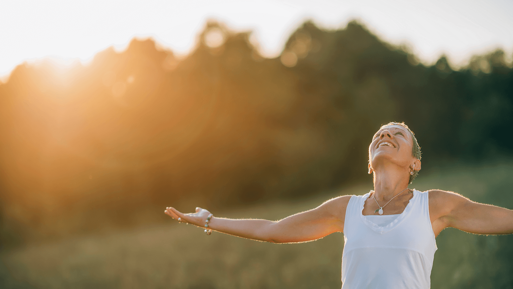 A mature woman stands in a sunlit field with her arms outstretched and a serene smile, embracing the warm golden light. This image represents the joy and freedom of living without pain, a new chapter of wellness supported by PRP's products.